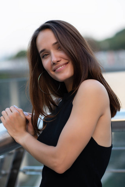Young pretty likable cheerful woman posing summer city outdoor in Kyiv, Ukraine. Beautiful self-confident girl with long brown hair posing on Dnipro river bank enjoing her life, urban lifestyle - Photo, image