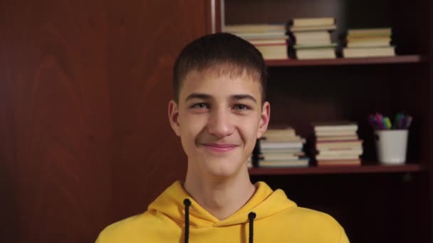 Young man student laughs and drinks from a white mug on the background of a bookcase with textbooks and school supplies. Close-up. Good morning mood on quarantine of corornavirus - Materiał filmowy, wideo