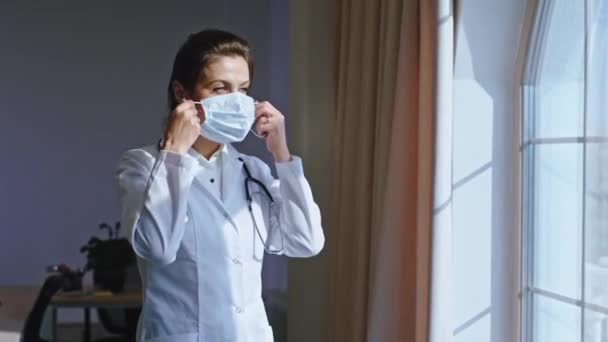 Charismatic mature woman doctor take her protective mask and wearing on her face and looking through the hospital panoramic window. - Imágenes, Vídeo