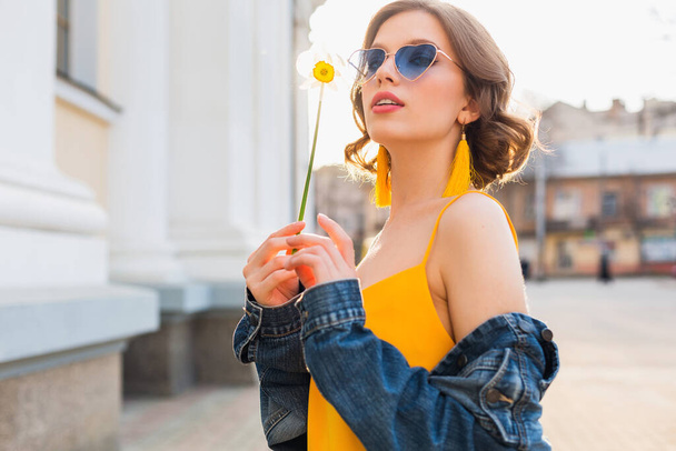 portrait of pretty woman wearing heart sunglasses holding flower against sun, sunny summer day, stylish apparel, fashion trend, blue jeans jacket, yellow dress, elegant hipster boho earrings - Fotó, kép