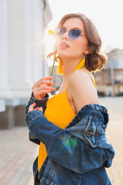bright portrait of beautiful woman holding flower, yellow dress, denim jacket, hipster style, summer fashion trend, smile, trendy sunglasses - 写真・画像