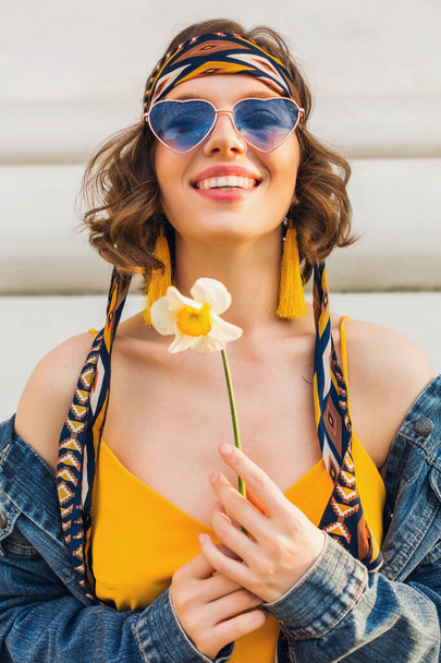 stylish attractive woman in hippie style outfit wearing yellow dress denim jacket, trendy accessories, sunglasses, smiling, holding flower, street fashion, spring summer fashion trend - Photo, image