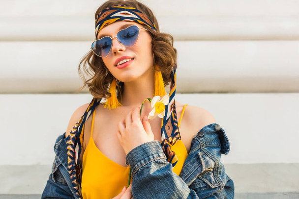 stylish attractive woman in hippie style outfit wearing yellow dress denim jacket, trendy accessories, sunglasses, smiling, holding flower, street fashion, spring summer fashion trend - Фото, изображение