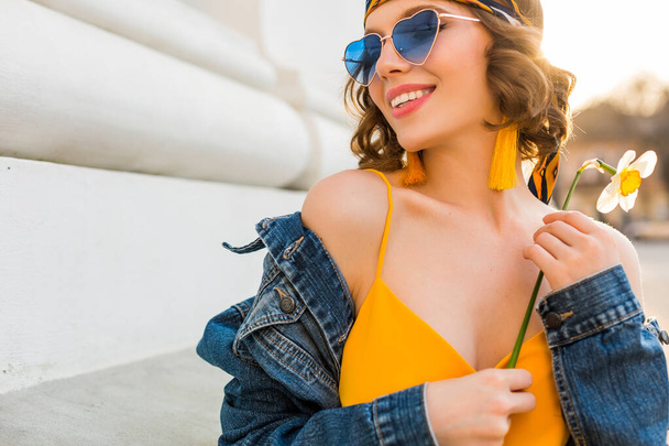 close up portrait of stylish attractive woman in hippie style outfit wearing yellow dress denim jacket, trendy accessories, sunglasses, smiling, holding flower, spring summer fashion trend - Photo, Image