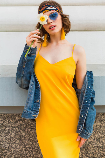 beautiful woman in yellow stylish dress wearing denim jacket, posing with flower, trendy outfit, hippie indie style, spring summer fashion trend, blue sunglasses, street fashion - Фото, зображення