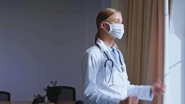 Senhora enfermeira atraente com uma máscara protetora fazer uma pausa ela tem um rosto pensando olhando através da janela panorâmica e tirar a máscara protetora Covid-19
 - Filmagem, Vídeo