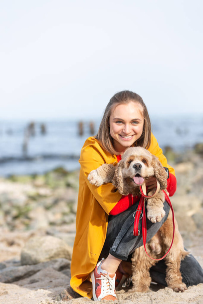 Egy sárga kabátos fiatal nő ölelget egy kutyát a tengerparton. A lány, aki engedelmes kutyát simogat a tengerparton. Legjobb barátok. - Fotó, kép