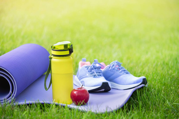 Zapatillas deportivas en estera de yoga y botella de agua, manzana sobre hierba verde fresca
. - Foto, Imagen