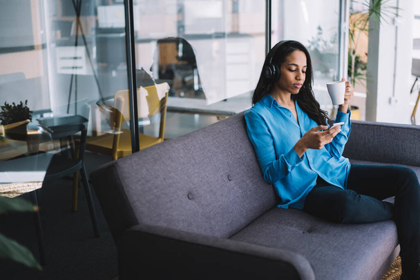 Chilling modern black woman in casual outfit sitting on couch and listening to music while browsing smartphone and drinking cup of coffee at home - Фото, изображение