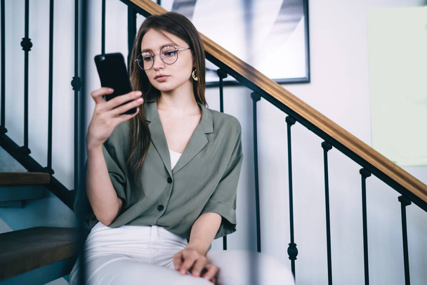 Focused serious slender woman in blouse and round glasses resting on contemporary decorative stairs and using smartphone in light interior - Fotoğraf, Görsel