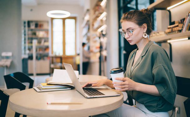 Young modern woman in casual outfit and with glasses sitting at table typing on laptop while drinking coffee against blurred shelves in library - Photo, Image