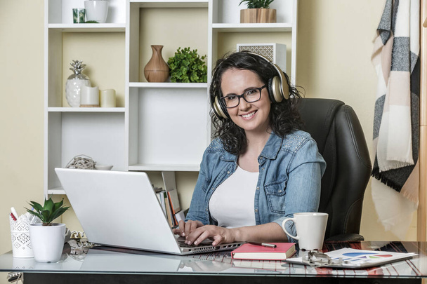Wesoła kobieta freelancer w okularach i słuchawkach bezprzewodowych w ubraniach casual uśmiecha się do kamery siedząc w miękkim skórzanym fotelu przy stole z kubkiem gorącego napoju i interakcji z netbook we współczesnym mieszkaniu - Zdjęcie, obraz