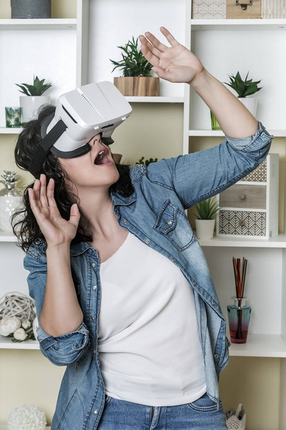 Erstaunte erwachsene Frau in Jeanskleidung gestikuliert und schreit, während sie die virtuelle Realität zu Hause erkundet - Foto, Bild