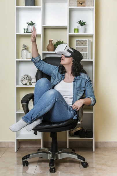 Pełna długość dorosła kobieta w drelichu i w okularach VR siedząca na krześle i interakcja z cyberprzestrzenią w domu - Zdjęcie, obraz