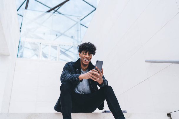 Laughing trendy young African American man in stylish clothing with curly hair using smartphone while sitting on stairs in white modern building - Fotó, kép