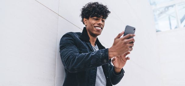 Happy curly black man in stylish denim suit with smartphone leaning in white wall while messaging in social media and smiling - 写真・画像