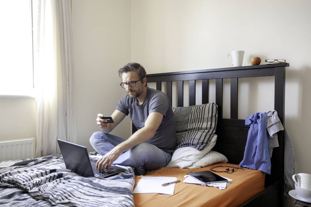 Man making online payment, sitting on bed while working from home
 - Фото, изображение