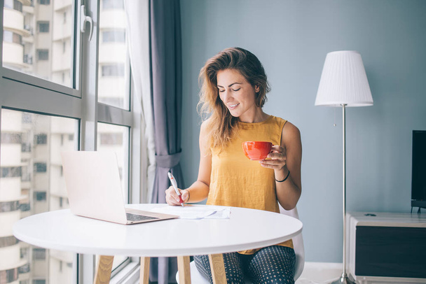 Joyful young slim woman in yellow sleeveless blouse sitting at white round table with laptop and red mug writing on paper at home - Valokuva, kuva