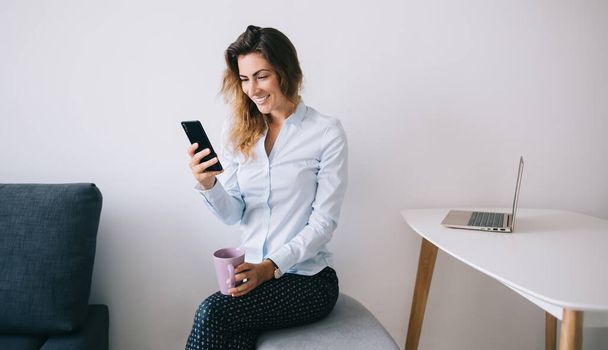Young female employee smiling and sitting with cup and legs crossed while texting on smartphone in office against white background - Foto, Imagem