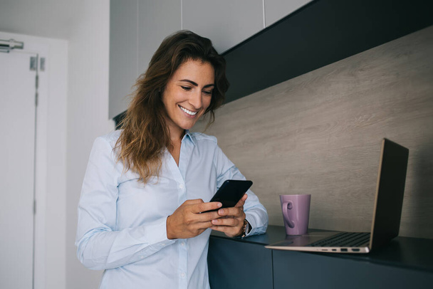 Piuttosto ottimista donna sorridente in camicia blu leggere buone notizie su smartphone mentre in piedi a casa vicino a laptop e tazza
 - Foto, immagini