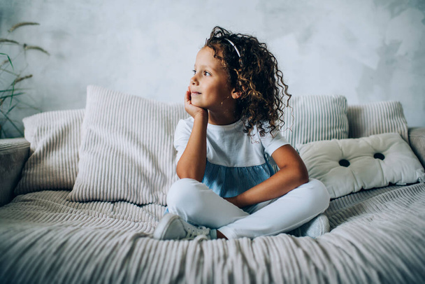Charming little curly girl in casual clothes sitting on comfy couch at home leaning on hand and looking away in dreams - Foto, immagini