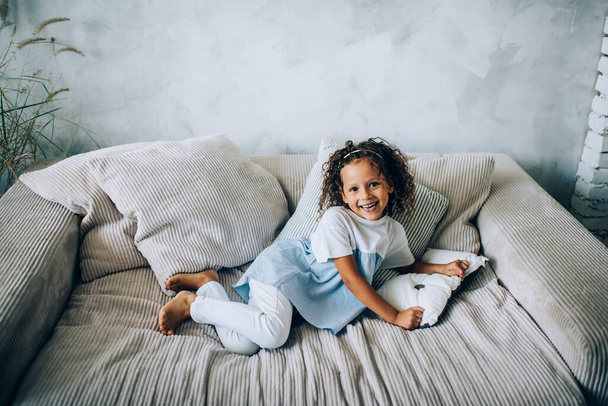 From above of laughing little curly girl in casual clothes lying on comfortable big couch at home smiling at camera - Foto, Bild