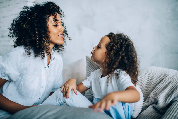 Charming alike ethnic mother and girl with curly hair sitting together on couch at home pouting lips in kiss and looking at each other - Zdjęcie, obraz