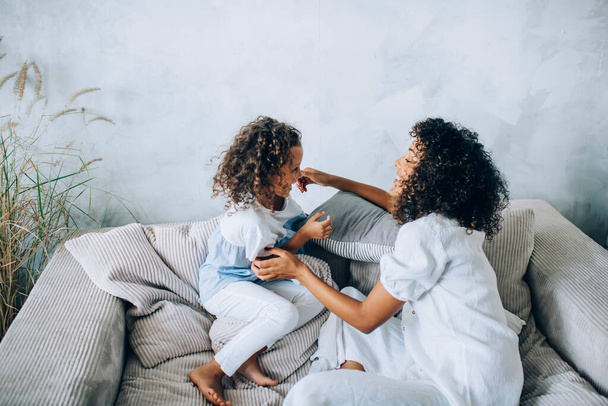 Side view of happy beautiful woman and girl both with curly hair having fun at home while chilling on comfy sofa - Photo, image