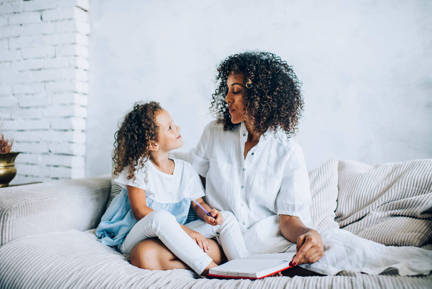 Beautiful ethnic woman and little daughter with curly hair sitting together on sofa with notepad and looking at each other with love - Fotografie, Obrázek