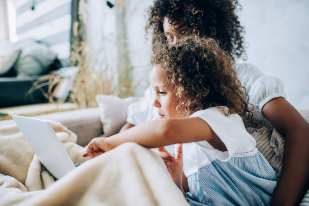 Side view of adorable little curly girl cuddling under plaid with mother sitting on couch at home and sharing laptop  - Foto, Imagen