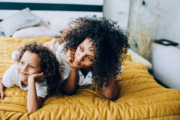 Charming cute little girl with mother both having curly hair lounging on bed at home leaning on hands and looking away in dreams - Fotoğraf, Görsel