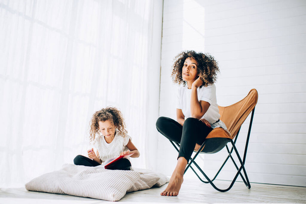 Young sensual curly woman sitting in armchair with adorable little daughter near writing in notepad and looking at camera in daylight - 写真・画像