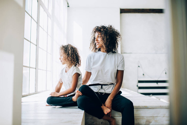 Beautiful ethnic curly woman and little girl wearing similar clothes sitting together in light white studio smiling away in window - Фото, изображение