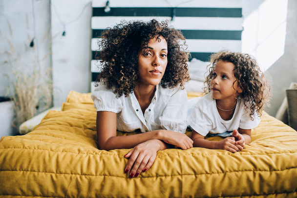 Wonderful ethnic curly woman with cute alike little girl lying together on bed at home spending time at home in leisure - Fotoğraf, Görsel