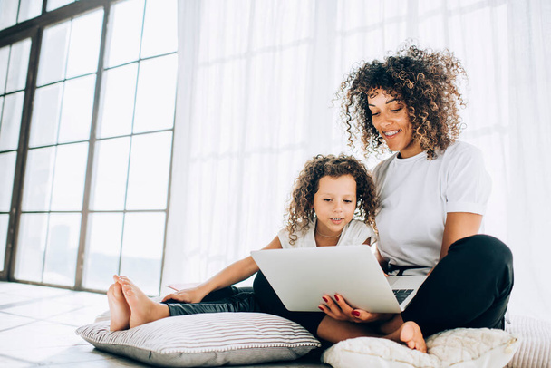 Lovely little adorable girl with beautiful curly mother sitting on cushions in bright daylight and watching laptop together smiling happily - Foto, imagen