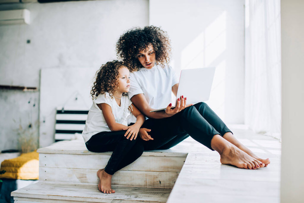 Beautiful ethnic curly woman with charming alike daughter sitting together in light room using modern laptop together while spending time at home - 写真・画像