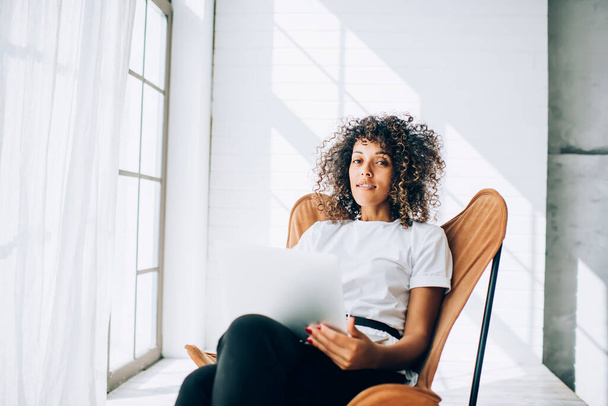 Young stylish ethnic woman with curls lounging in armchair in light white room holding modern laptop on knees while looking at camera - Zdjęcie, obraz