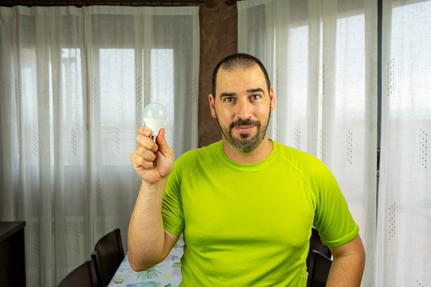 Uomo con barba e capelli corti in camicia verde felice con una lampadina in salotto a casa. Idea concetto
 - Foto, immagini