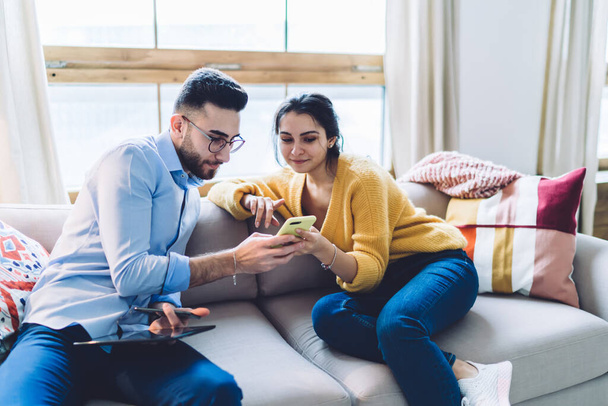 Young ethnic casual male in shirt and glasses and female in sweater sitting on sofa at home and searching information in mobile phone - Foto, Bild