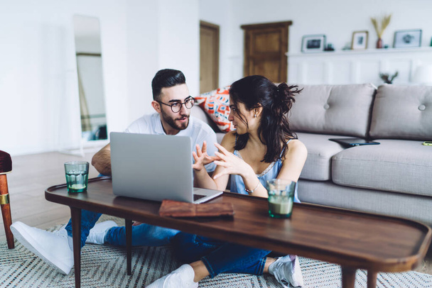 Couple sitting together on floor at table with laptop while woman gesticulating and sharing ideas with listening man in glasses on background with sofa - Photo, Image