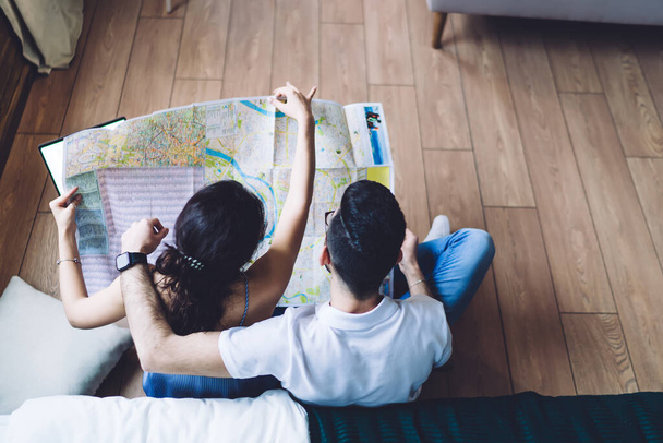Overhead back view of romantic couple relaxing on wooden floor looking down at paper map and dreaming about vacation at home - Foto, Bild