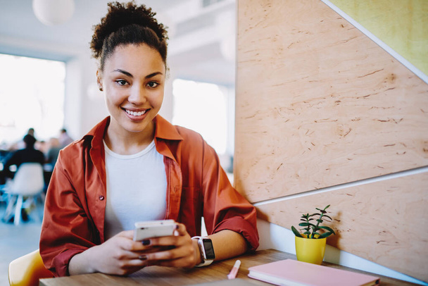 African female blogger with cute smile on face looking at camera while chatting with friend via mobile application for communication,positive hipster girl sitting at cafeteria table and using cellular - Φωτογραφία, εικόνα