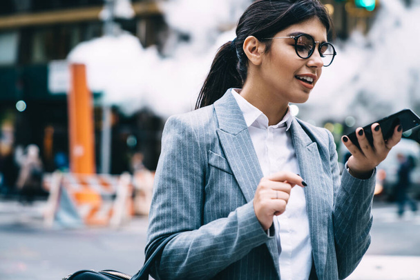 Happy young female entrepreneur in round glasses and stylish classic suit walking at street with smartphone and speaking on phone - Фото, изображение