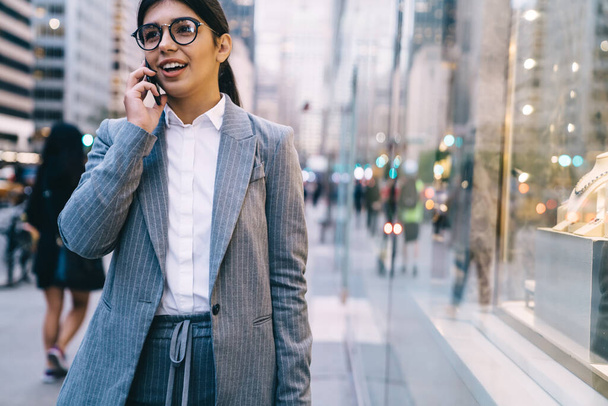 Crop of smiling beautiful adult employee female in formal outfit and eyeglasses having phone call while walking on street on background of blurry people looking away  - Fotó, kép