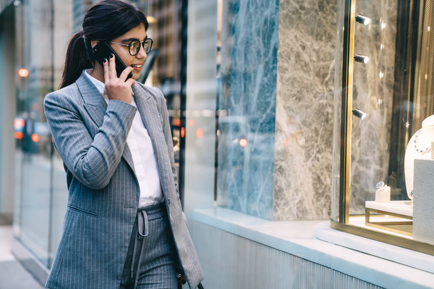 Confident pretty adult worker female in formal outfit and eyeglasses and with black hair talking on smartphone while walking on street on background of blurry people looking at shop  - Foto, afbeelding