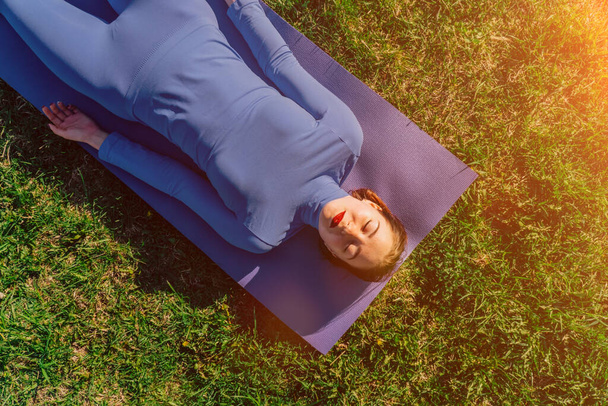 A young Caucasian girl is standing in a pose on a mat on the grass during sunset. The concept of exercise for peace of mind. Fitness stretching - Photo, image