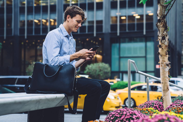 Cheerful handsome young man in classic outfit sitting hand leaning on leather bag texting on smartphone front of commercial building on street with cars of New York city  - Foto, Imagem