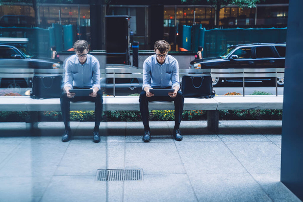 Przystojny, skupiony młodzieniec w okularach siedzący na ławce odbijający w szklanej ścianie używając tabletu na ulicy na Manhattanie, Nowy Jork - Zdjęcie, obraz