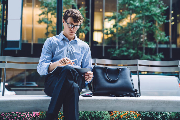 Handsome concentrated young male in glasses sitting on bench in front of office building browsing tablet on street in Manhattan, New York city - Foto, afbeelding
