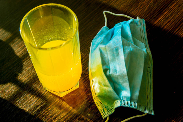 Glass of fizzy yellow vitamins drink near the medical blue mask. Illness prevention and health care concept - Fotografie, Obrázek